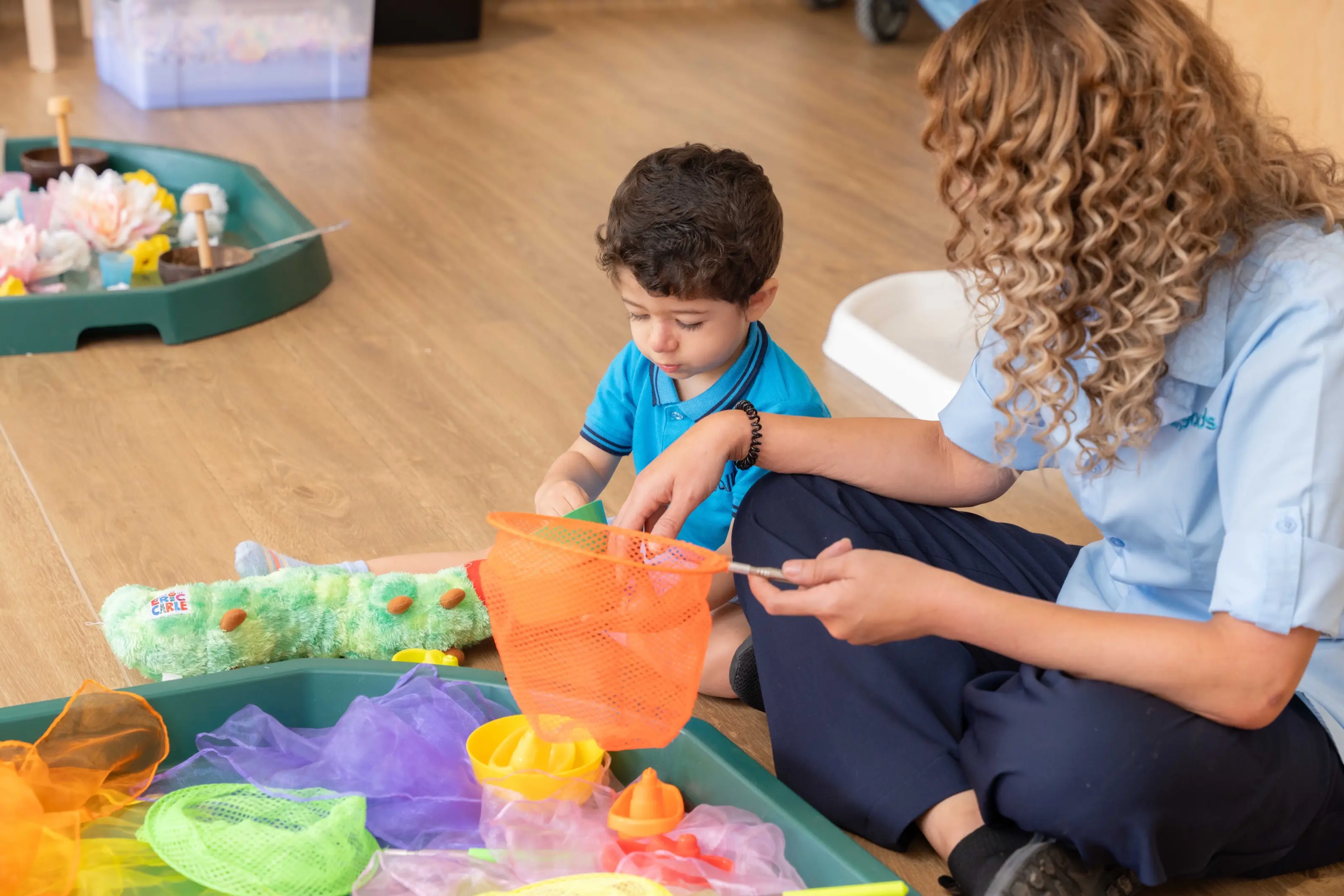 A child at Royal Kids Nursery lifting a toy from a sensory tray with an orange net, an educator kneeling beside him